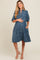 woman wearing a denim blue maternity dress with short puff sleeves, button front, and side pockets, showcasing her baby bump