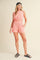 woman wearing a pink two-piece set with a crop top and shorts, paired with a black cap and slide sandals
