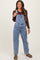 woman wearing a light blue maternity denim overall with a brown long sleeve top and sandals, holding the straps