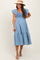 woman wearing a light blue maternity dress with ruffle sleeves and a smocked bodice, standing with a hat and sandals