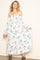 woman wearing a white off-the-shoulder floral maxi dress with blue and orange flowers, posing with one hand on her hip