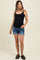 woman wearing a black maternity tank top and denim shorts, showing her baby bump with a casual smile.