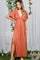 woman wearing a rust-colored maxi dress with a deep v-neck and side slits, standing beside a white chair and floral backdrop