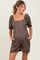 woman wearing a brown checkered maternity romper with short puff sleeves and pockets, showing her baby bump