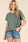woman wearing an olive green ribbed short sleeve top paired with distressed denim shorts, smiling at the camera