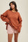 woman wearing an oversized rust orange knit sweater and light pink shorts, poses with one hand on her hip