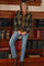 woman wearing a green plaid maternity jacket over a brown top and blue jeans, posing with one foot on a shelf