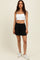 woman wearing a white cropped tank top and black athletic shorts, standing against a plain background