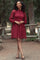 woman wearing a maroon long sleeve smocked dress with a ruffled hemline, posing outdoors among trees