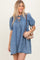 woman wearing a light denim short sleeve dress with a tie neckline and puff sleeves, holding a small brown bag