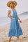 woman wearing a light blue tiered maxi dress with pockets and a white tee, standing on the beach with a straw hat