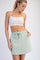 woman wearing a light green drawstring skirt and a white cropped tank top, posing with one hand on her hip