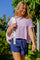 woman wearing a light purple cropped t-shirt and navy blue shorts, walking outdoors with a backpack over her shoulder