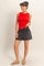 woman wearing a red sleeveless button-up top and black smocked waist shorts, standing with one hand in pocket