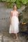 woman wearing a white sundress with red floral embroidery and spaghetti straps, standing in a garden setting