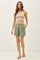 woman wearing a light pink spaghetti strap crop top and green pleated shorts, standing with one hand in her pocket
