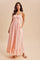 woman wearing a light pink maxi dress with adjustable straps and ruffle hem, standing with hands in pockets