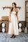 woman wearing a cream maxi dress with thin straps and black detailing, standing in a bright room with a patterned rug