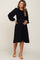 smiling pregnant woman wearing a black long sleeve smocked maternity midi dress and tan heels