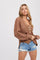 Woman wearing a brown chunky knit button-up cardigan and distressed denim shorts against a white background