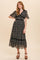 woman wearing a black floral patterned maxi dress with short flutter sleeves and a tiered skirt, standing on a light background