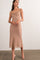 woman wearing a tan midi dress with a draped neckline and slim straps, standing in a relaxed pose