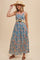 woman wearing a blue floral maxi dress with a belted waist and a straw hat, smiling and posing with one hand raised