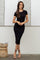 woman wearing a black ribbed midi dress with short sleeves and strappy high heels, standing against a minimalist background