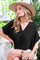 woman wearing a black loose-fitting v-neck top with wide sleeves and a straw hat, seated with a clutch bag