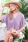 woman wearing a lavender oversized blouse with a v-neck and holding a woven clutch while sitting outdoors in a sun hat