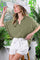 woman wearing an olive green oversized button-up shirt and white shorts, standing in a bright greenery-filled setting