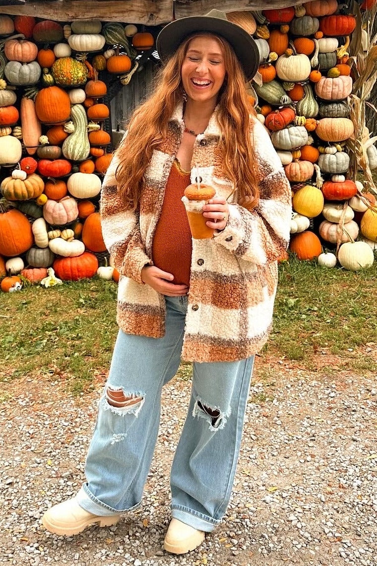 woman wearing a tan and white plaid maternity shacket, holding a drink, with visible baby bump and distressed jeans