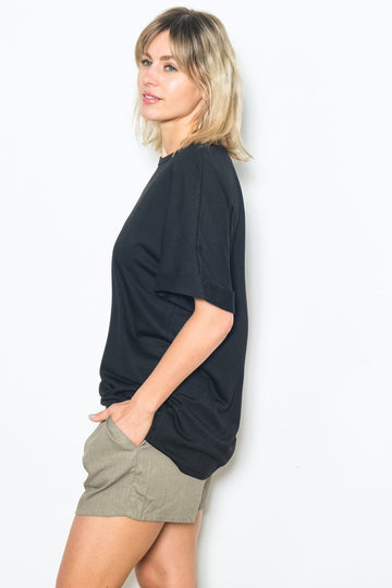 woman wearing a black oversized t-shirt and beige shorts, standing sideways with hands in pockets