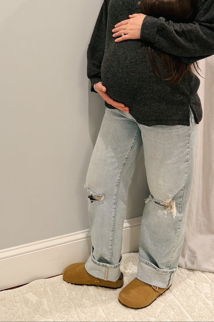 woman wearing a dark gray sweater and distressed light blue maternity jeans, cradling her baby bump