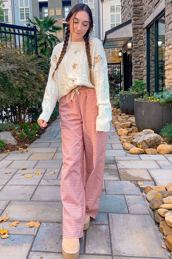 woman wearing a cream cable knit sweater with floral details and pink checkered wide-leg pants, standing outdoors