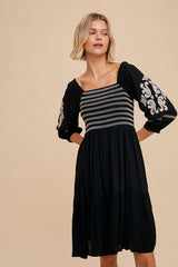 woman wearing a black dress with white embroidered detail and puff sleeves, standing against a neutral background