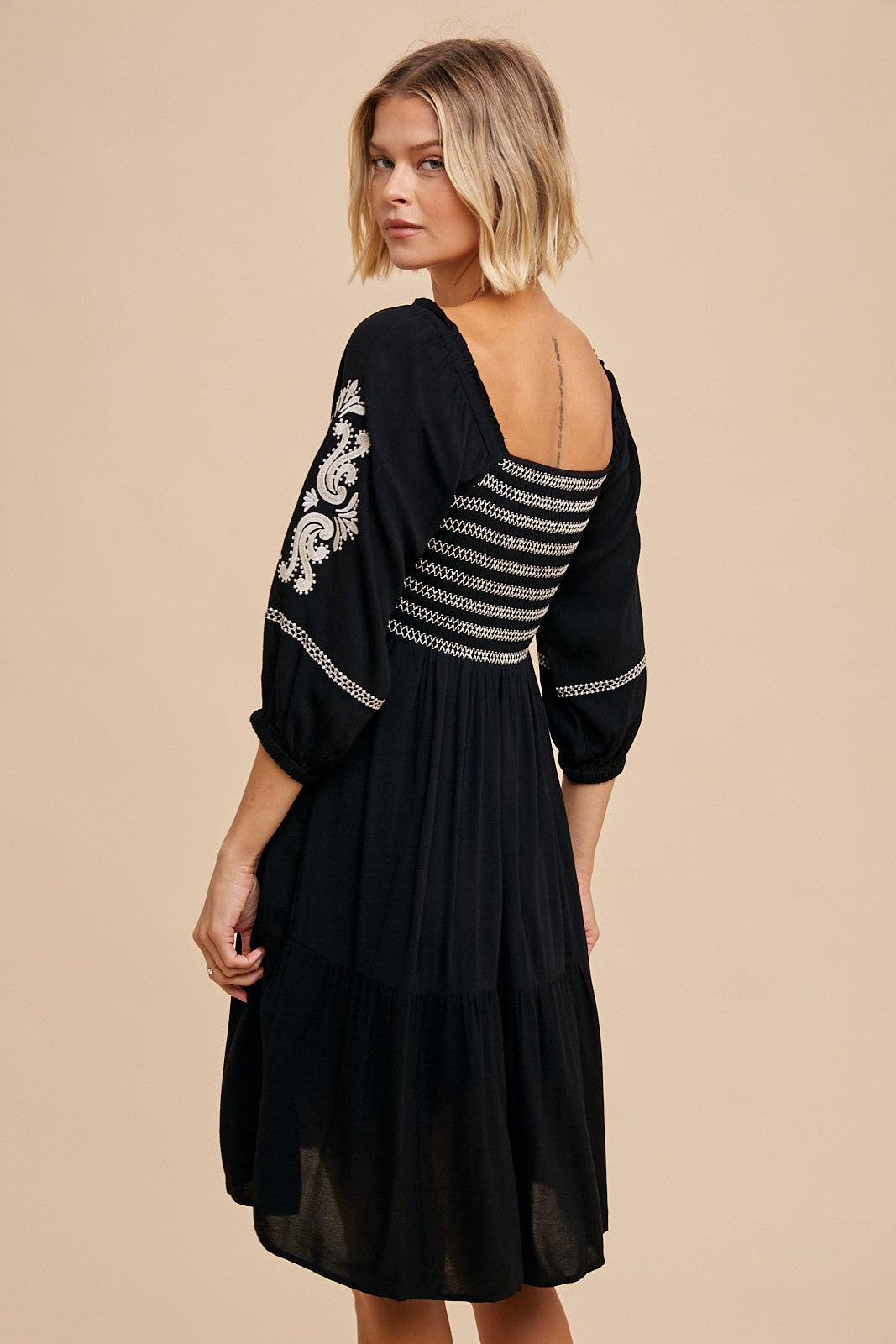 woman wearing a black embroidered dress with three-quarter sleeves and a tiered skirt, viewed from the back