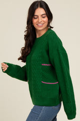 woman wearing a green cable knit sweater with pink stripe detailing and side pockets, styled with blue jeans