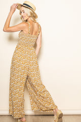 woman wearing a mustard floral maxi jumpsuit with smocked bodice and shoulder straps, facing away and wearing a straw hat
