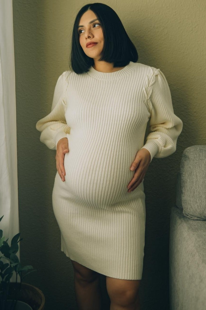 woman wearing a cream ribbed maternity dress, showcasing her baby bump while posing by a light wall.