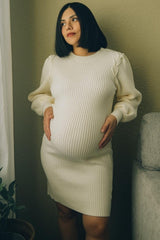 woman wearing a cream ribbed maternity dress, showcasing her baby bump while posing by a light wall.
