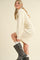 woman wearing a cream ribbed sweater dress with puff sleeves and knee-high black boots, smiling and posing sideways