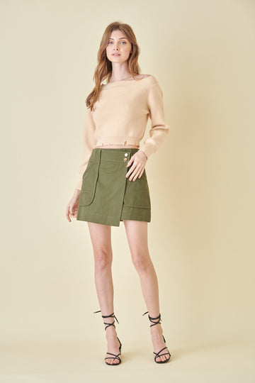 woman wearing a beige cropped sweater and an olive green mini skirt with front pockets and button details