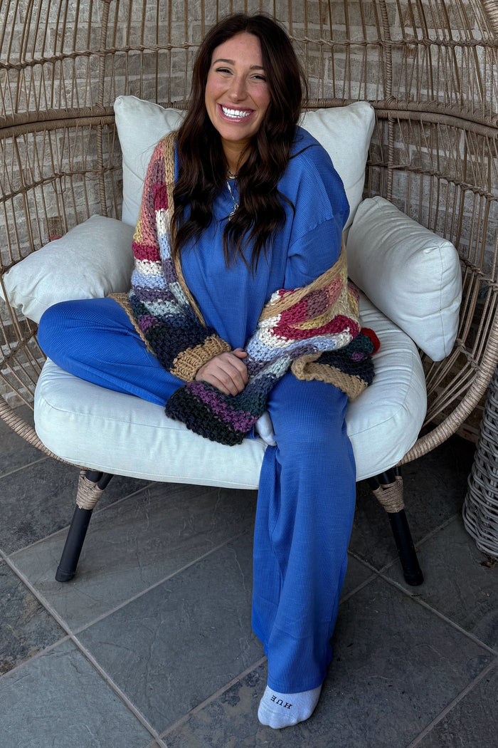woman wearing a blue loungewear set with a multicolored chunky knit cardigan, sitting on a chair with a smile