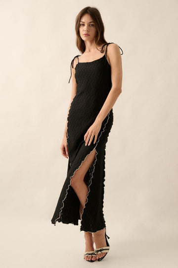 woman wearing a black maxi dress with tie shoulder straps and scalloped edges, featuring a thigh-high slit