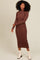 woman wearing a brown long sleeve ribbed midi dress and white ankle boots, smiling with hands on her hips