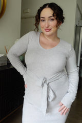 woman wearing a light grey ribbed maternity dress with a tie detail at the waist and long sleeves, showing her baby bump