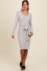 woman wearing a light gray long sleeve sweater dress with a tie waist detail and side slit, standing and smiling