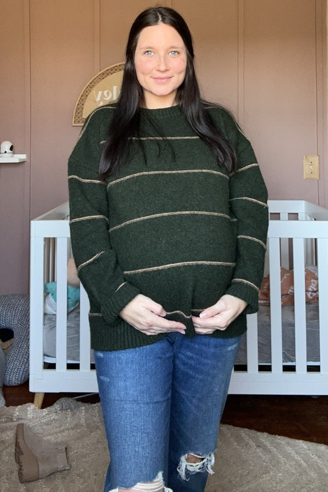 woman wearing a dark green striped maternity sweater and showing her baby bump while standing in front of a crib