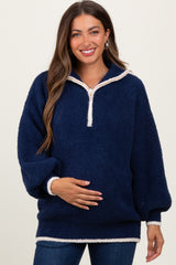 woman wearing a navy blue fuzzy maternity pullover with a quarter zip and white trim, holding her baby bump